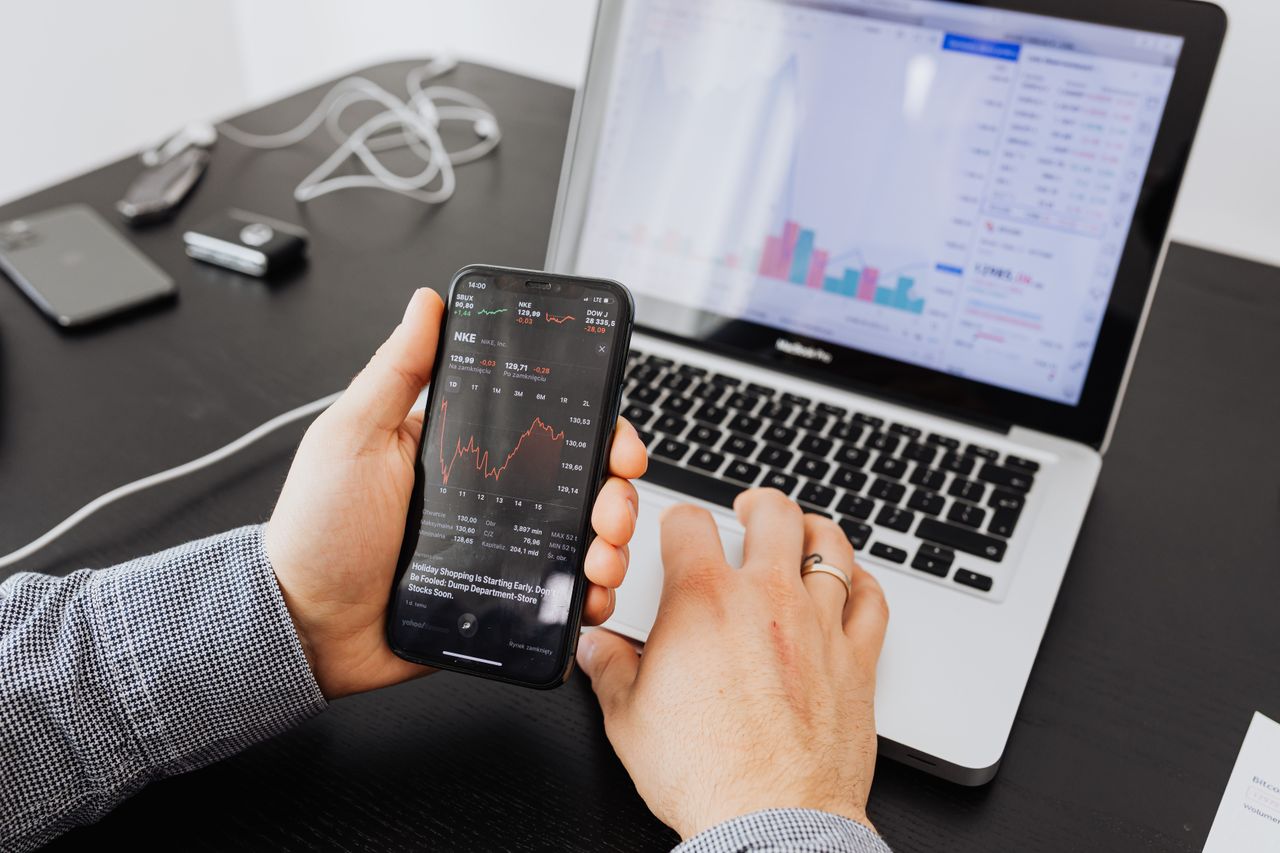 Une personne analyse des données boursières avec un smartphone et un ordinateur portable, en intérieur.
